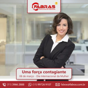 Uma mulher aparece de braços cruzados e sorrindo para a câmera. Ela está em um ambiente de trabalho e veste roupas de escritório.